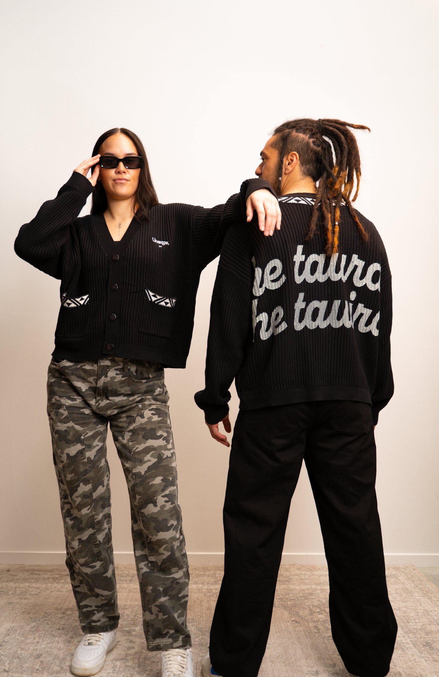 Two people wearing black jackets with text, one in front and one in back, on a plain background.