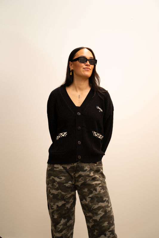 Person wearing a black cardigan and camouflage pants against a plain background